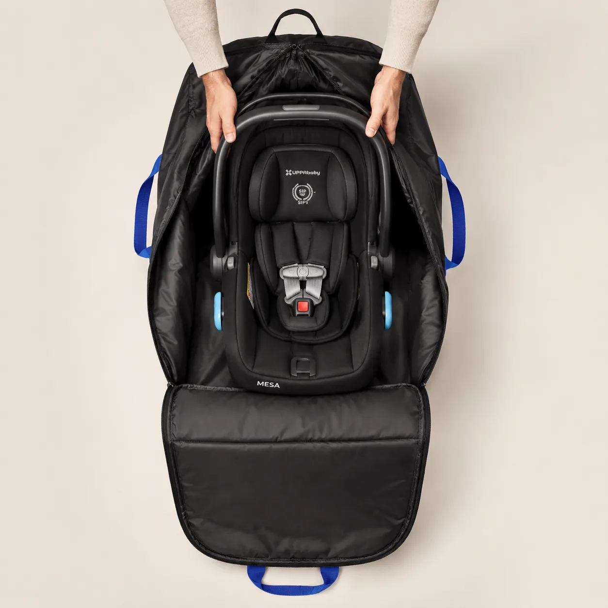 Overhead view of two hands holding open the padded black SeatPorter travel bag with blue carry straps, an infant car seat carrier sitting inside on the lining