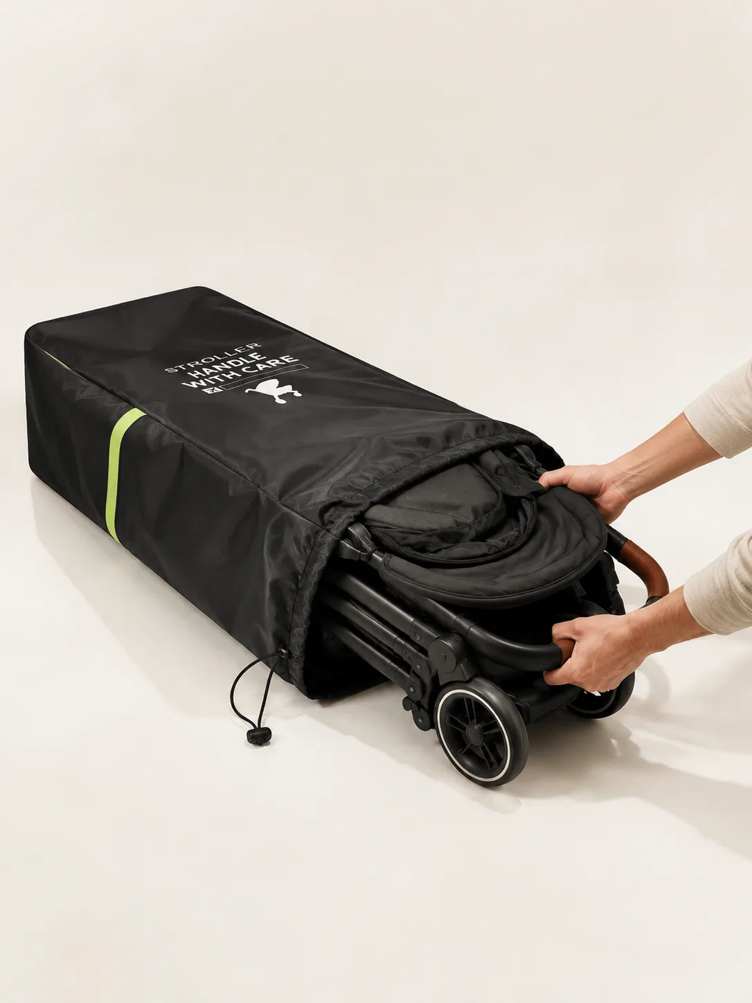 Two hands sliding a folded stroller with its wheels attached into the black stroller travel bag lying flat, drawstring cord hanging loose and a green reflective band across the side